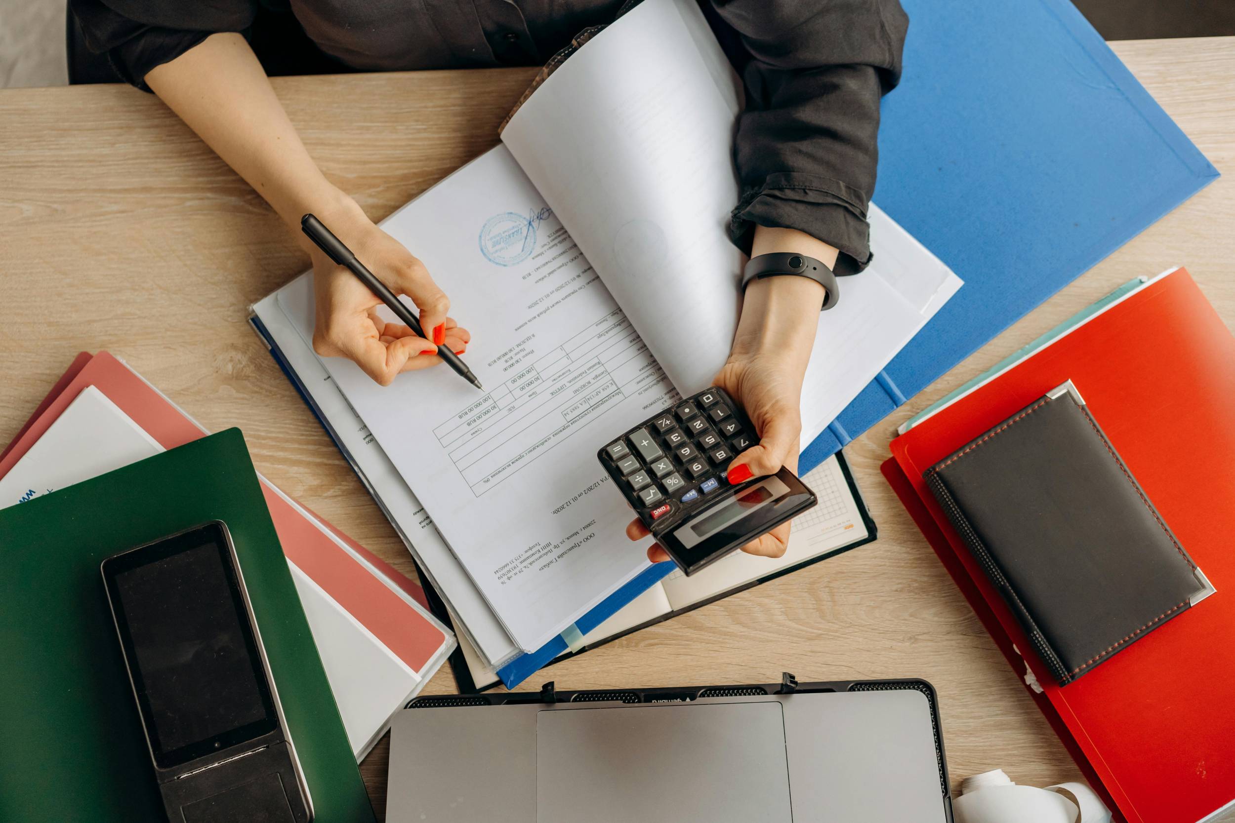 Overhead view of a person checking a document with a pen and calculator among colorful file folders