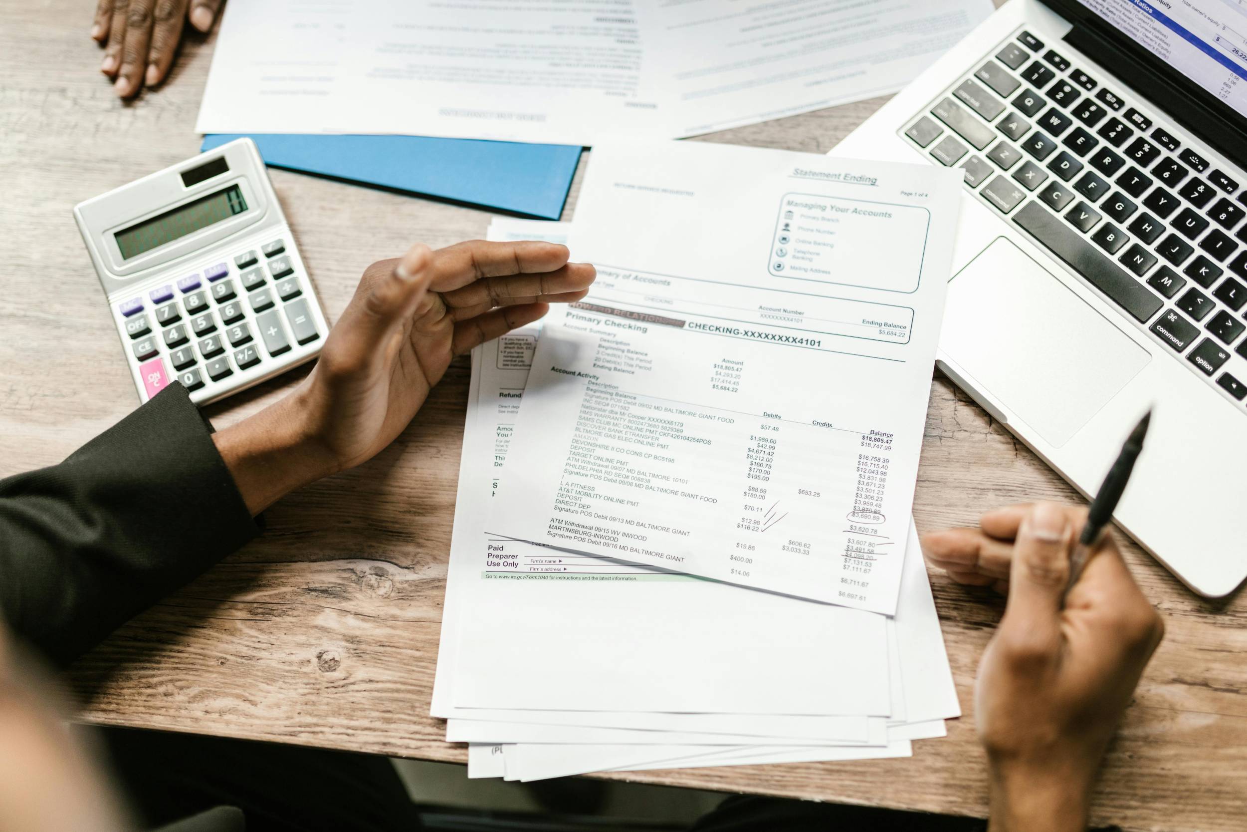 Hands reviewing a bank statement and tax documents beside a calculator and laptop